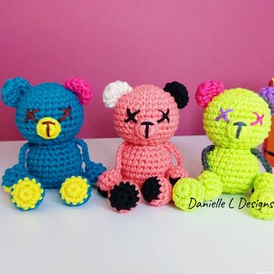 May include: Three crocheted teddy bears with stitched-on eyes and mouths. The bears are sitting on a white surface. The bear on the left is blue with yellow feet, the middle bear is pink with black feet, and the bear on the right is green with yellow feet. The text "Danielle L Designs" is visible in the bottom right corner of the image.