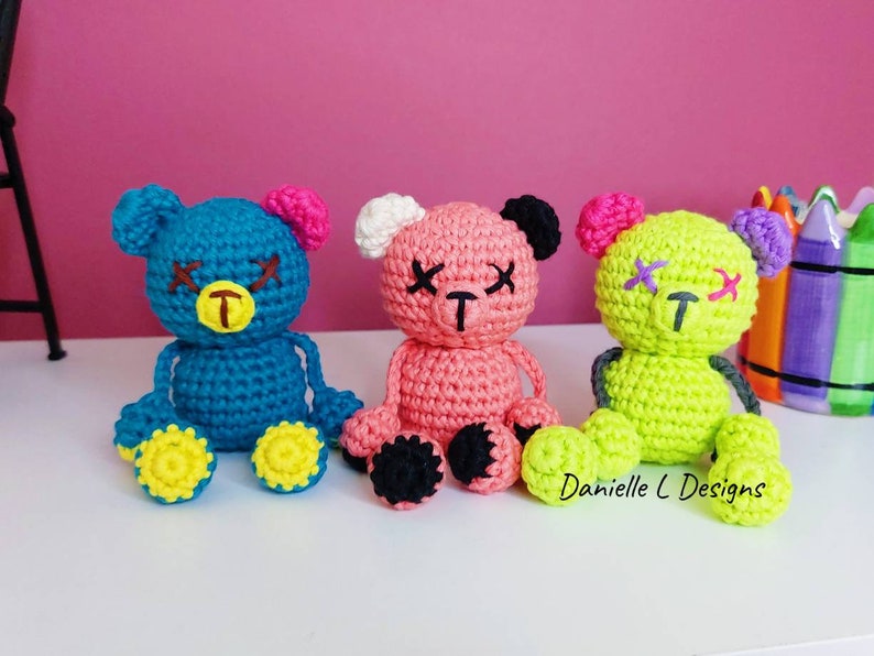 May include: Three crocheted teddy bears with stitched-on eyes and mouths. The bears are sitting on a white surface. The bear on the left is blue with yellow feet, the middle bear is pink with black feet, and the bear on the right is green with yellow feet. The text "Danielle L Designs" is visible in the bottom right corner of the image.