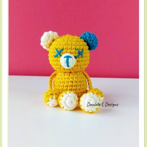 May include: A yellow crocheted teddy bear with white feet and blue ears. The bear has embroidered eyes and a nose with the letter "T" on its chest. The bear is sitting on a white surface with a pink background.