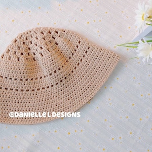May include: A crocheted beige bucket hat with a delicate openwork pattern. The hat is lying on a white background with a floral pattern.