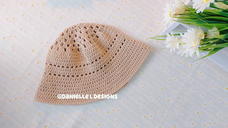 May include: A crocheted beige bucket hat with a delicate openwork pattern. The hat is lying on a white background with a floral pattern.