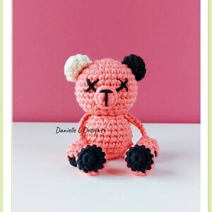 May include: A pink crocheted teddy bear with black eyes and black feet. The bear has white ears and a black nose.