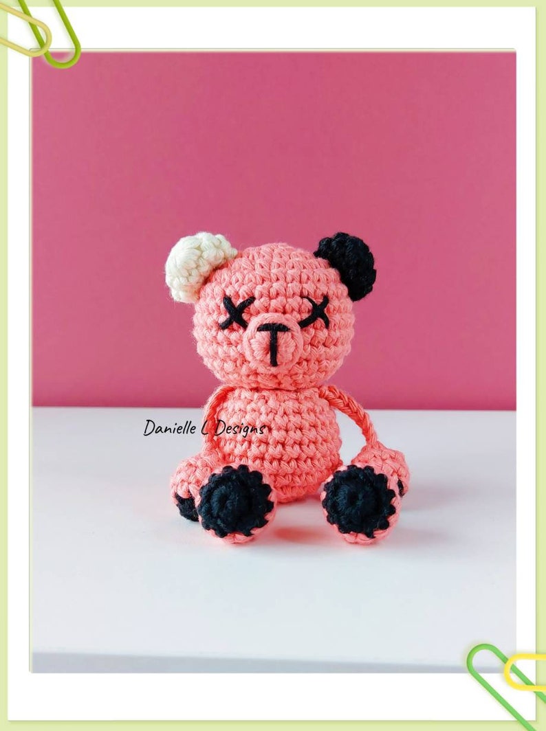 May include: A pink crocheted teddy bear with black eyes and black feet. The bear has white ears and a black nose.