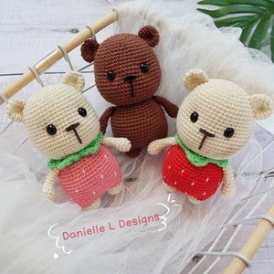 May include: Three crocheted teddy bears, two are cream colored with red and green strawberry outfits and one is brown with no outfit. The bears are sitting in a wooden basket.