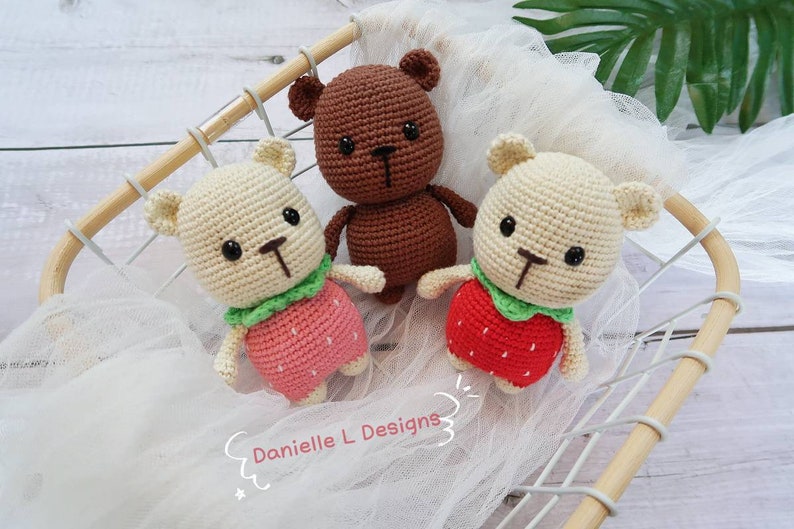 May include: Three crocheted teddy bears, two are cream colored with red and green strawberry outfits and one is brown with no outfit. The bears are sitting in a wooden basket.
