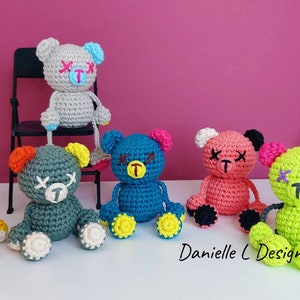 May include: Six crocheted teddy bears with stitched-on eyes and mouths. The bears are in various colors, including yellow, gray, teal, pink, and green. The bears are sitting on a white surface.  Danielle C Designs.