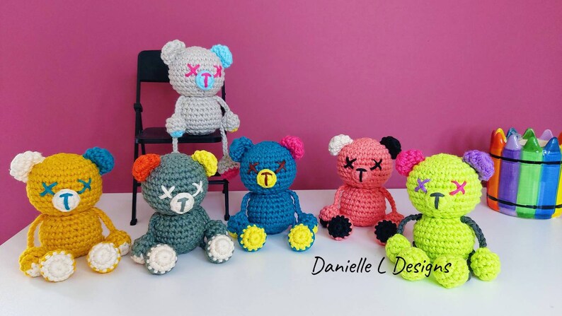 May include: Six crocheted teddy bears with stitched-on eyes and mouths. The bears are in various colors, including yellow, gray, teal, pink, and green. The bears are sitting on a white surface.  Danielle C Designs.