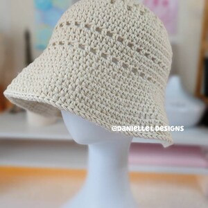 May include: A crocheted sun hat in a cream color with a wide brim and an open weave pattern. The hat is displayed on a white mannequin.