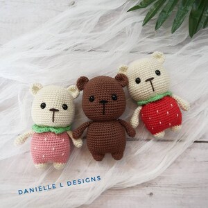 May include: Three crocheted stuffed bears, one brown and two white, wearing strawberry-themed outfits. The bears are sitting on a white fabric background.