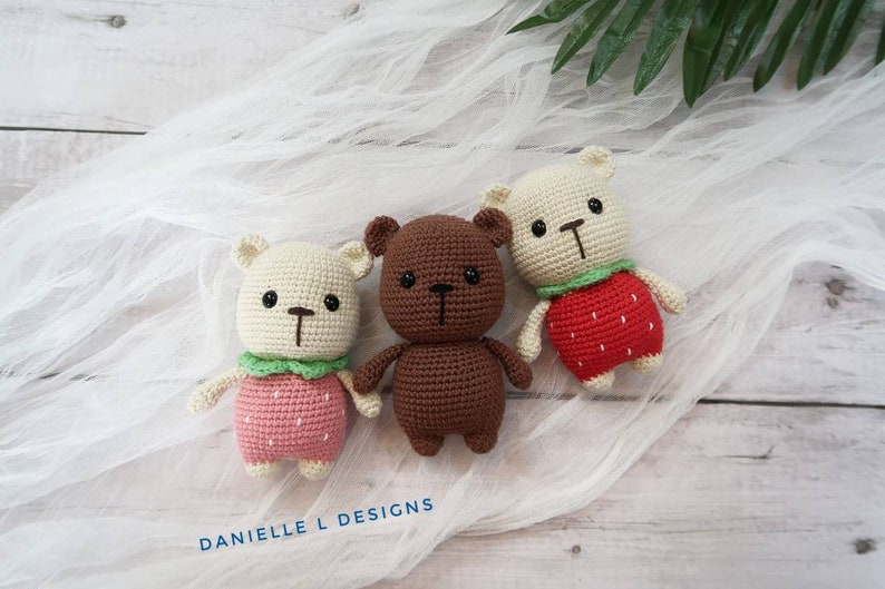 May include: Three crocheted stuffed bears, one brown and two white, wearing strawberry-themed outfits. The bears are sitting on a white fabric background.