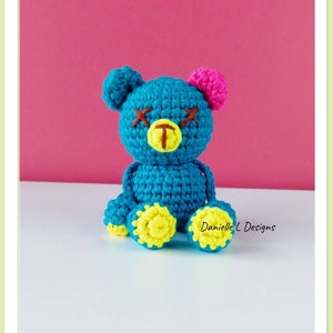 May include: A crocheted blue teddy bear with yellow feet, a pink ear, and brown stitched eyes. The bear is sitting on a white surface.  Danielle L Designs.