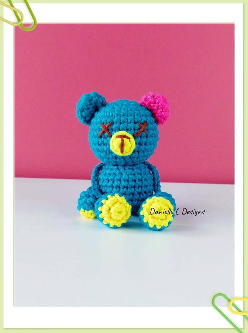 May include: A crocheted blue teddy bear with yellow feet, a pink ear, and brown stitched eyes. The bear is sitting on a white surface.  Danielle L Designs.