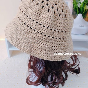 May include: A beige crocheted bucket hat with a wide brim and an open weave pattern. The hat is sitting on a mannequin with brown curly hair.