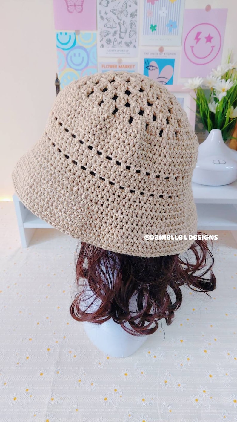 May include: A beige crocheted bucket hat with a wide brim and an open weave pattern. The hat is sitting on a mannequin with brown curly hair.