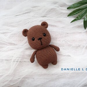 May include: A brown crocheted teddy bear with black eyes and a black nose. The bear is lying on a white fabric background.