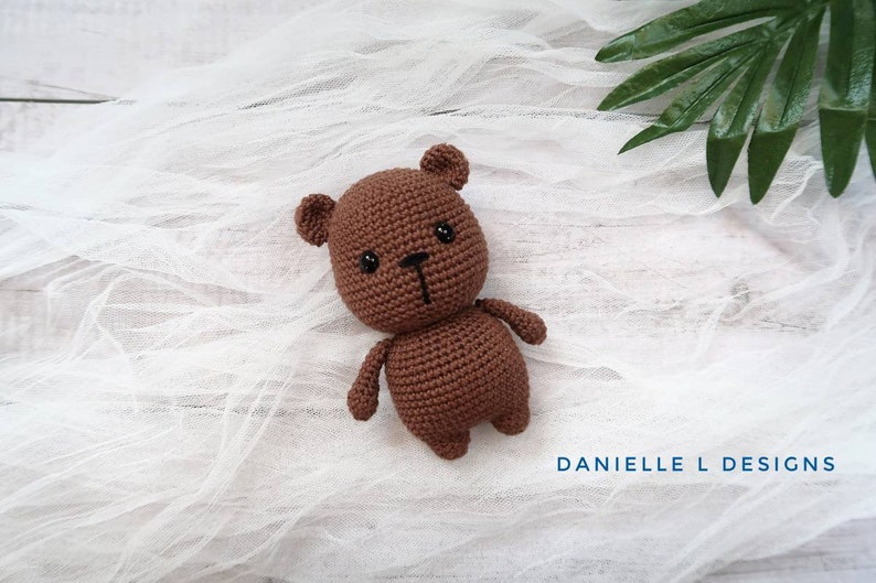 May include: A brown crocheted teddy bear with black eyes and a black nose. The bear is lying on a white fabric background.