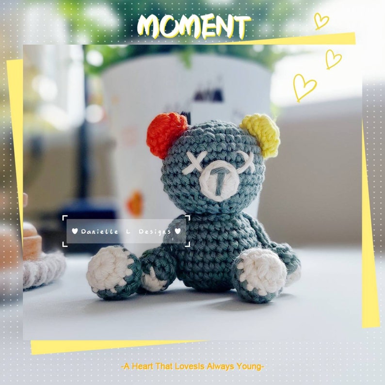 May include: A crocheted plush toy bear with a blue-grey body, white accents, and a yellow and red ear. The bear has a white face with black eyes and a stitched-on letter 'T'. The bear is sitting on a wooden surface. The text 'Danielle L Designs' is visible in the image.