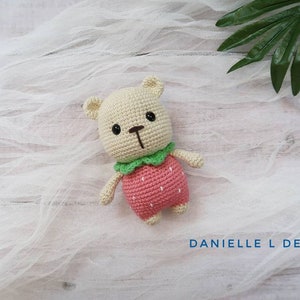 May include: A crocheted stuffed animal bear wearing a pink strawberry costume with green leaves. The bear is sitting on a white fabric background.