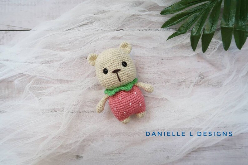 May include: A crocheted stuffed animal bear wearing a pink strawberry costume with green leaves. The bear is sitting on a white fabric background.
