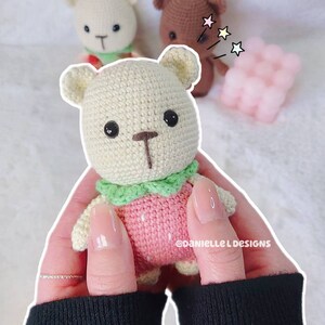 May include: A crocheted strawberry-themed teddy bear with a green collar, held in a person's hands. The bear is light brown with black eyes and a brown nose. The background is a white fuzzy surface.