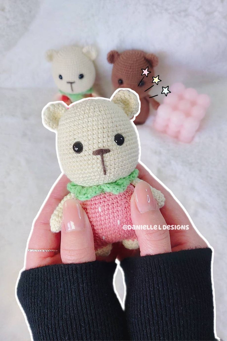 May include: A crocheted strawberry-themed teddy bear with a green collar, held in a person's hands. The bear is light brown with black eyes and a brown nose. The background is a white fuzzy surface.