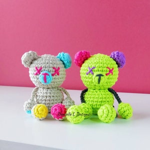 May include: Two crocheted teddy bears with stitched-on eyes and mouths. The bear on the left is gray with blue and pink accents. The bear on the right is green with purple and pink accents. Both bears have black yarn for arms and legs.