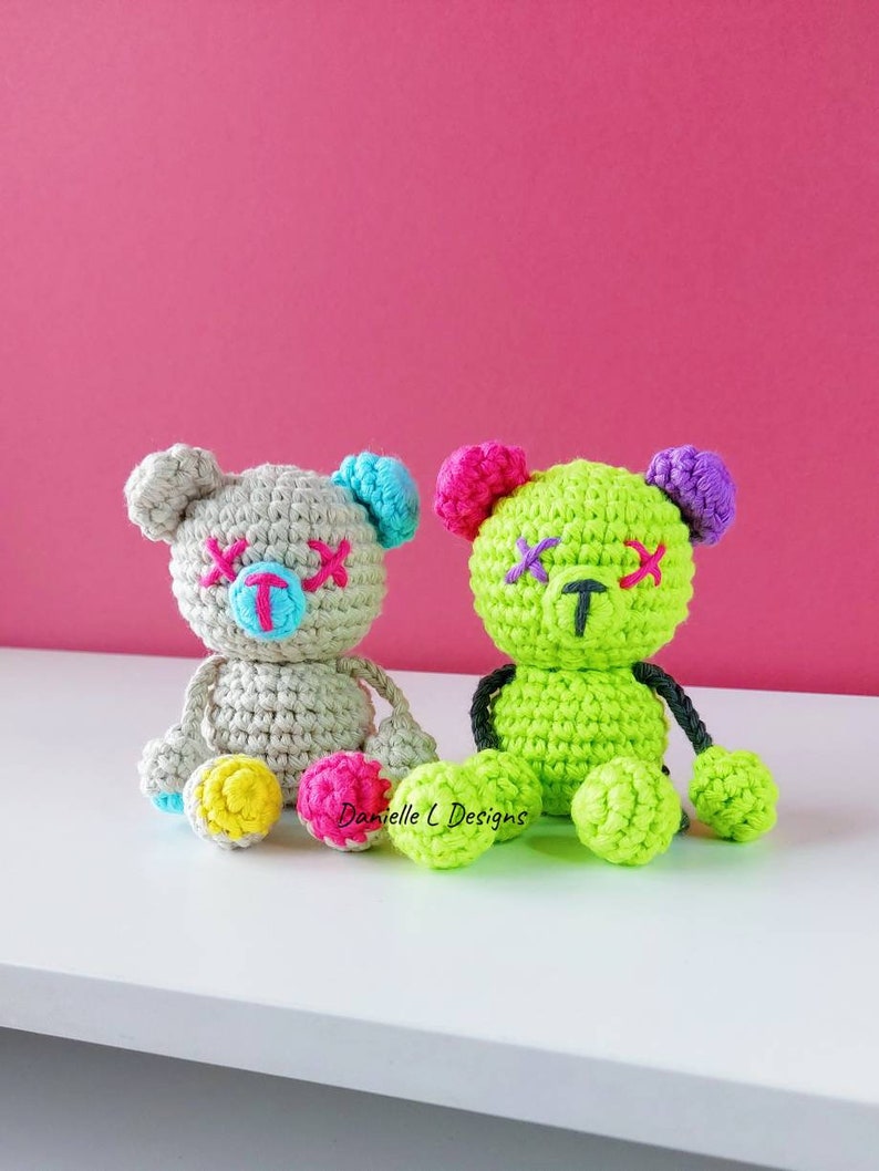 May include: Two crocheted teddy bears with stitched-on eyes and mouths. The bear on the left is gray with blue and pink accents. The bear on the right is green with purple and pink accents. Both bears have black yarn for arms and legs.