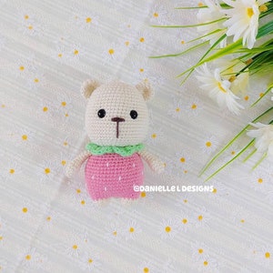 May include: A crocheted stuffed bear wearing a pink strawberry dress with green leaves. The bear has brown eyes and a brown nose. The bear is sitting on a white background with a floral pattern.