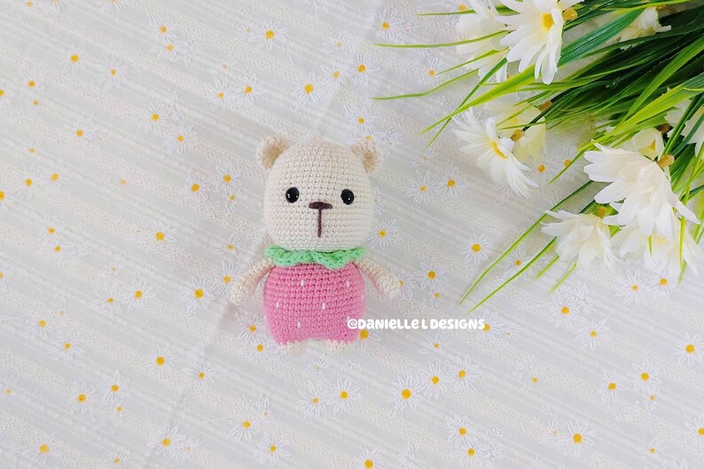 May include: A crocheted stuffed bear wearing a pink strawberry dress with green leaves. The bear has brown eyes and a brown nose. The bear is sitting on a white background with a floral pattern.