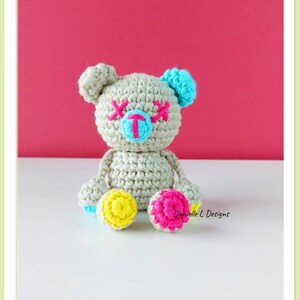 May include: A crocheted teddy bear with a light gray body, blue ears, pink eyes, and a blue nose. The bear has yellow and pink accents on its feet.  Danielle L Designs.