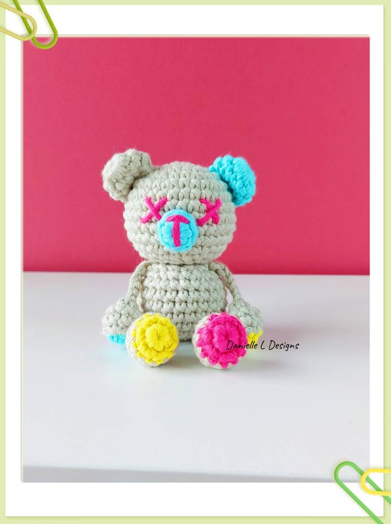 May include: A crocheted teddy bear with a light gray body, blue ears, pink eyes, and a blue nose. The bear has yellow and pink accents on its feet.  Danielle L Designs.