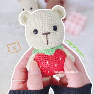 May include: A crocheted stuffed bear wearing a red strawberry costume with green leaves. The bear has black eyes and a brown nose.