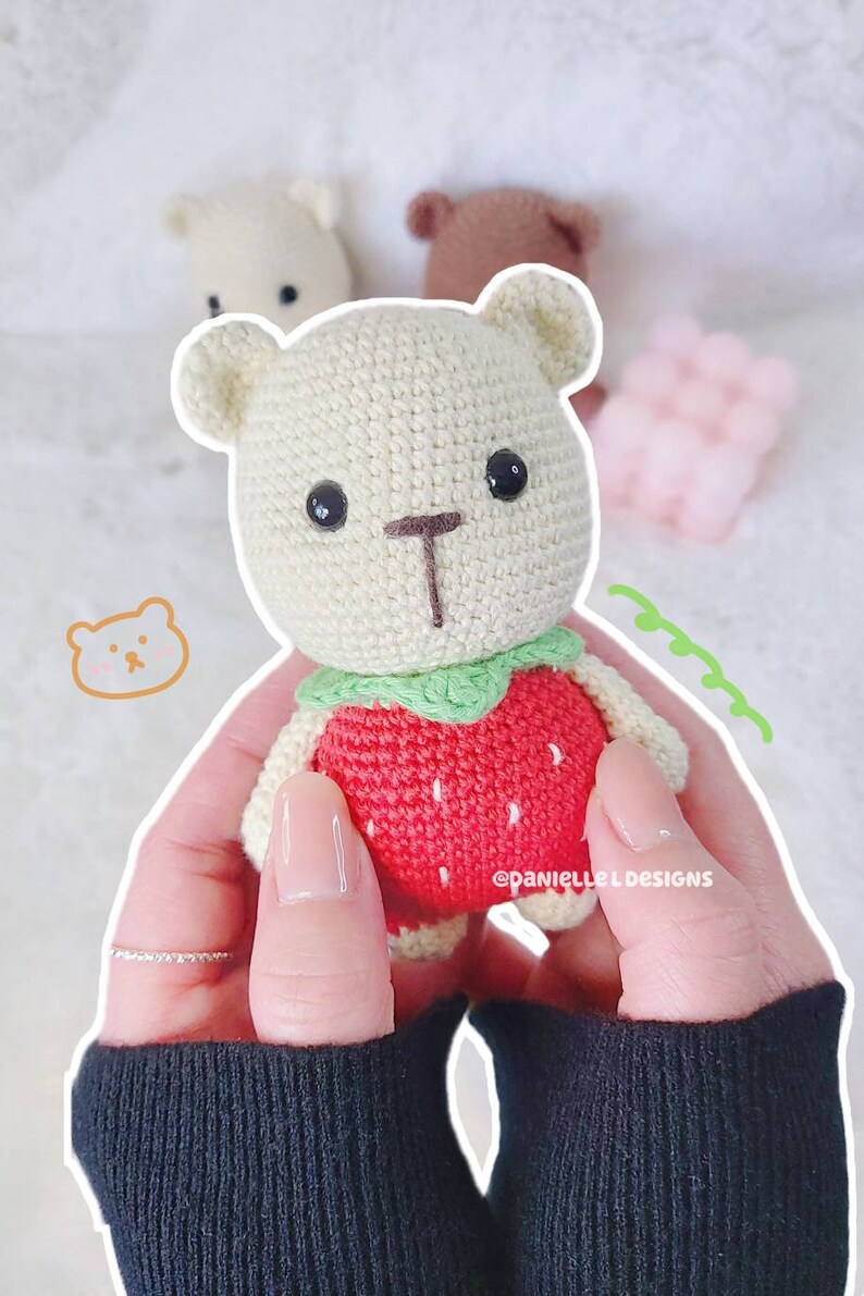 May include: A crocheted stuffed bear wearing a red strawberry costume with green leaves. The bear has black eyes and a brown nose.