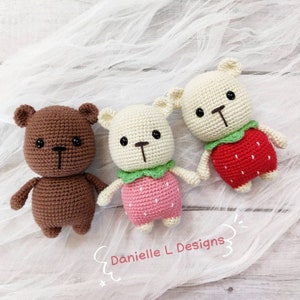 May include: Three crocheted stuffed bears wearing strawberry costumes. The bears are brown, white, and white. The strawberries are red with green leaves. The bears are sitting on a white fabric background.