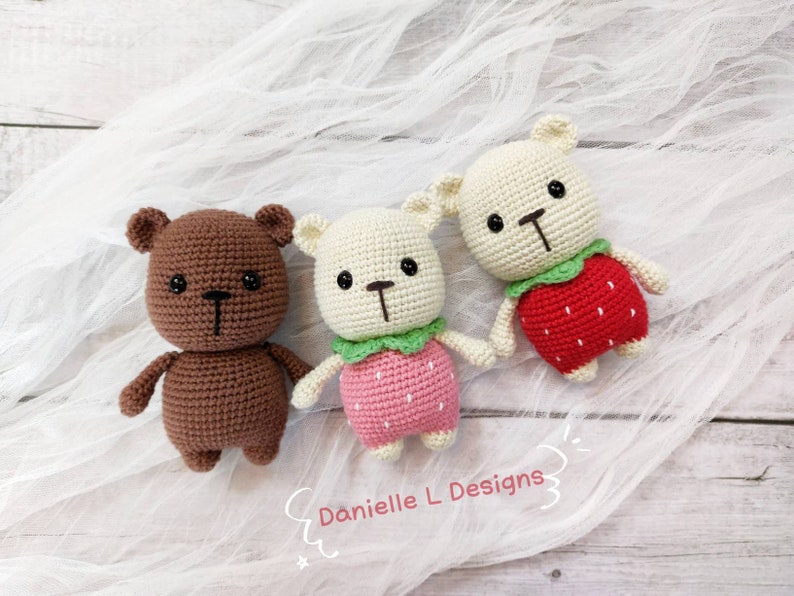 May include: Three crocheted stuffed bears wearing strawberry costumes. The bears are brown, white, and white. The strawberries are red with green leaves. The bears are sitting on a white fabric background.