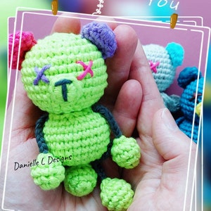 May include: A green crocheted teddy bear with pink, purple, and blue accents. The bear has stitched X's for eyes and a T for a nose. The bear is being held in a person's hands.