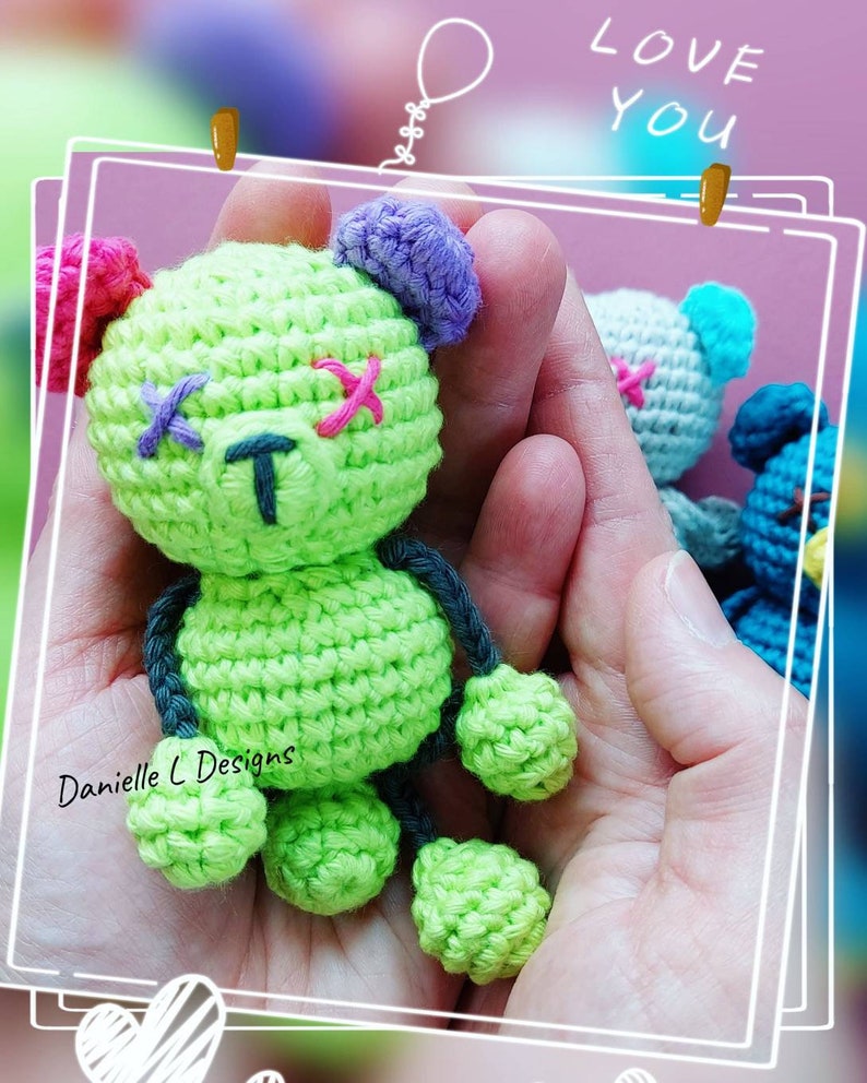 May include: A green crocheted teddy bear with pink, purple, and blue accents. The bear has stitched X's for eyes and a T for a nose. The bear is being held in a person's hands.