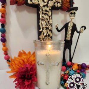 May include: A white candle with a silver cross design in a clear glass jar. The candle is lit and there is a small flame visible. The candle is surrounded by colorful flowers and a skeleton figurine.