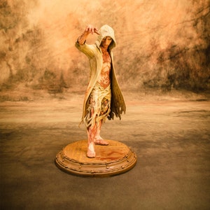 May include: A detailed figurine of a hooded figure with a pale, bloody, and skeletal appearance. The figure is standing on a circular base with a barbed wire border.