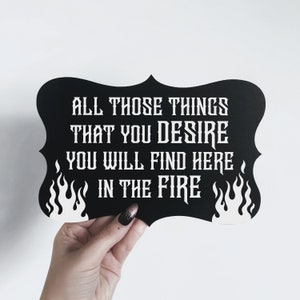 May include: Black sign with white text and flames on the sides. The text reads "All those things that you desire you will find here in the fire."