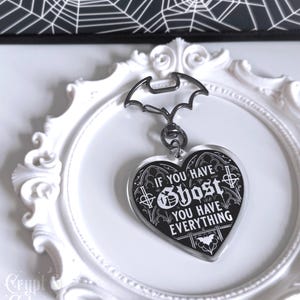 Puede incluir: Un llavero en forma de corazón con el texto "If You Have Ghost You Have Everything" en blanco sobre fondo negro. El llavero tiene un cierre en forma de murciélago y se muestra en un marco decorativo blanco.