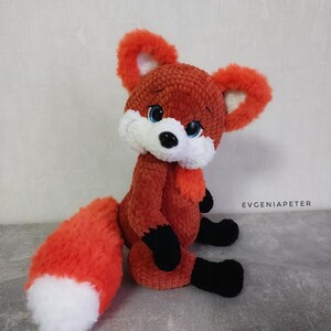 Könnte beinhalten: Ein gehäkeltes Plüschtier in Form eines Fuchses mit einem rot-weißen Schwanz, schwarzen Pfoten und blauen Augen. Der Fuchs sitzt auf einer grauen Oberfläche.