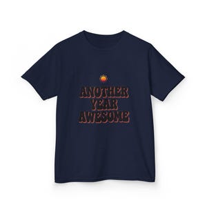 Peut inclure: Un t-shirt bleu marine à manches courtes avec une petite icône de soleil orange au-dessus du texte "ANOTHER YEAR AWESOME" imprimé dans une police sombre et rétro avec un léger contour orange, centré sur la poitrine du vêtement.