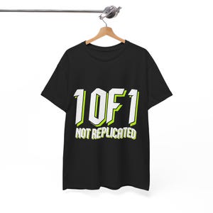 Könnte beinhalten: Ein schwarzes Kurzarm-T-Shirt mit dem weißen Text 1 OF 1 mit lindgrüner Umrandung und dem Text NOT REPLICATED in Weiß mit lindgrüner Umrandung darunter, präsentiert auf einem Holzkleiderbügel vor einem schlichten weißen Hintergrund.