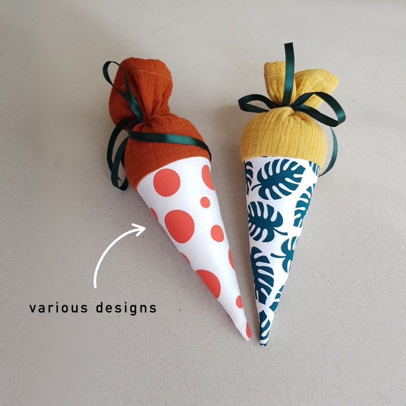 May include: Two fabric cones with different designs. One cone is orange and white with red polka dots. The other cone is yellow and white with a green tropical leaf pattern. The cones are sewn shut at the top and have a ribbon tied around the middle.