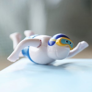 May include: A white plastic toy swimmer with blue and yellow goggles, lying on a light blue surface.
