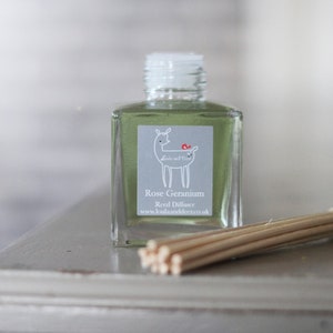 Puede incluir: Una botella de vidrio transparente con un líquido verde en su interior. La botella tiene una etiqueta blanca con el texto "Rose Geranium Reed Diffuser" y una pequeña ilustración de un ciervo. La botella está sobre una superficie marrón claro con un paquete de palitos de madera.