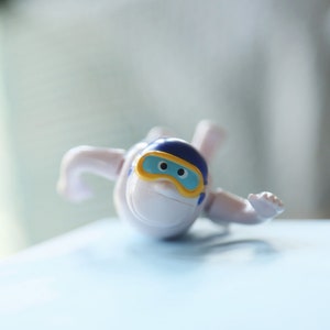 May include: A white plastic toy figure wearing a blue and yellow helmet with a blue face mask. The figure is lying on its stomach with its arms outstretched.