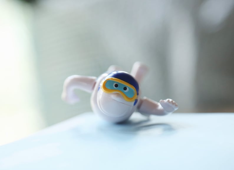 May include: A white plastic toy figure wearing a blue and yellow helmet with a blue face mask. The figure is lying on its stomach with its arms outstretched.