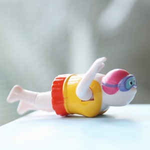 May include: A yellow, red, and white wind-up swimmer toy with a pink and blue swimming mask. The toy is in a swimming position with its arms outstretched.